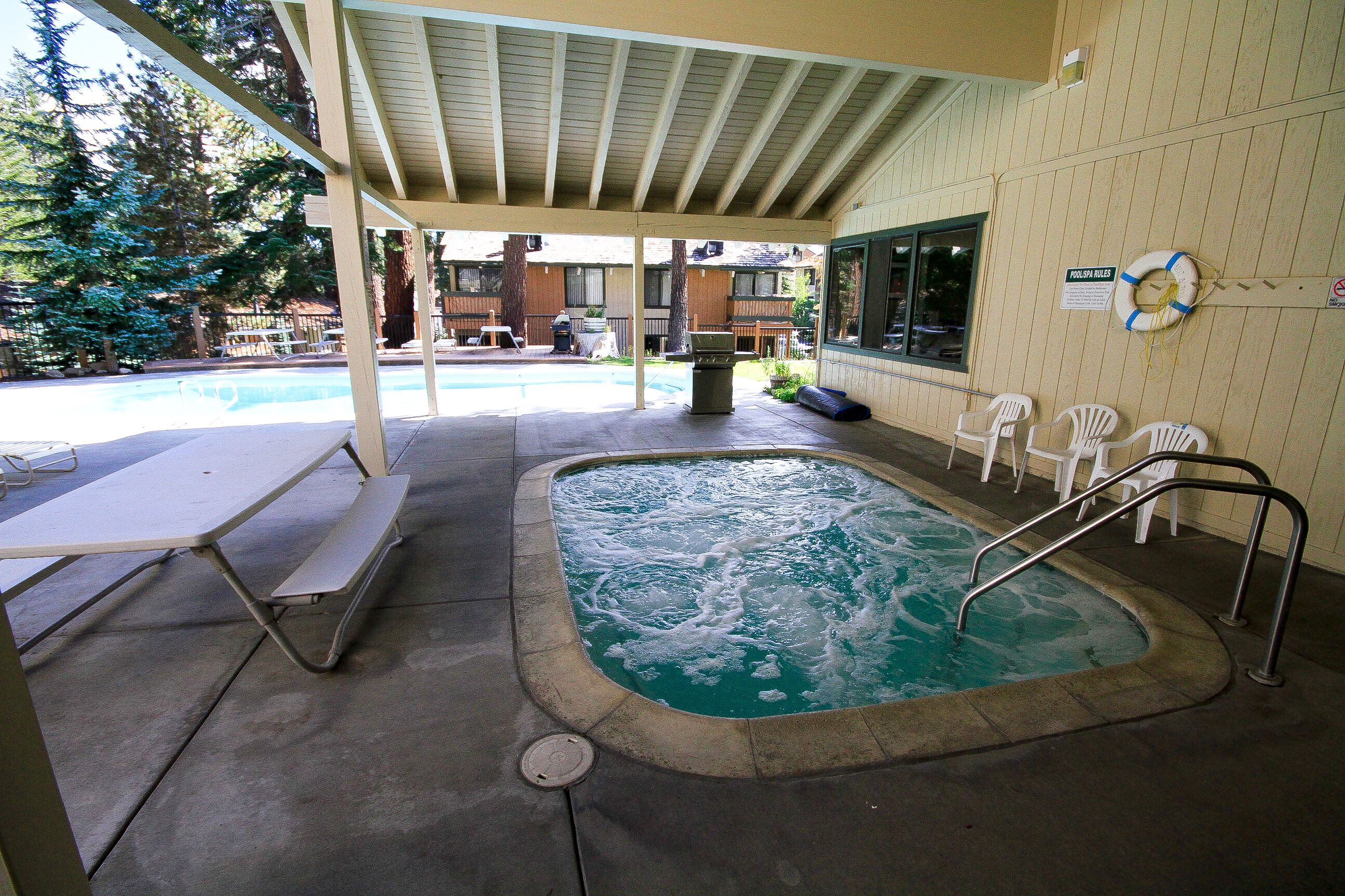 A covered outdoor pool area with a hot tub, picnic table, and white chairs.