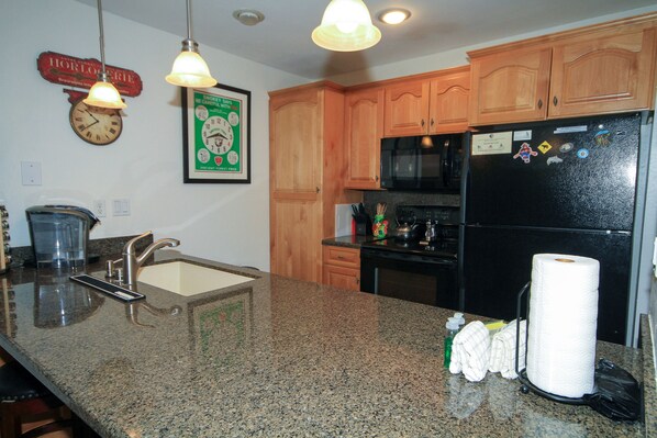 A kitchen with granite countertops, wooden cabinets, a stainless steel refrigerator, and a clock on the wall.