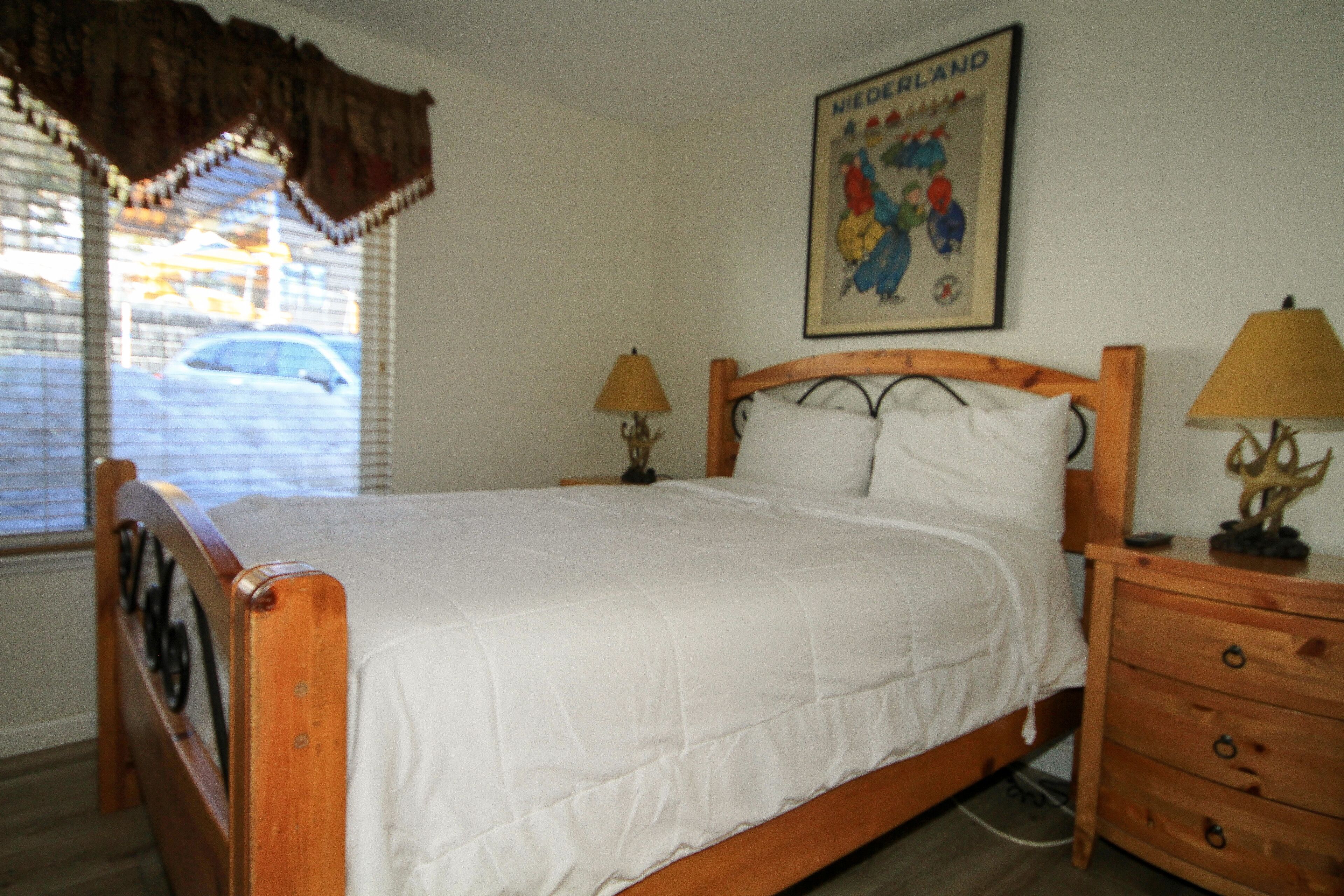A bedroom with a wooden bed, two bedside tables with lamps, a window with blinds, and a wall-mounted poster.