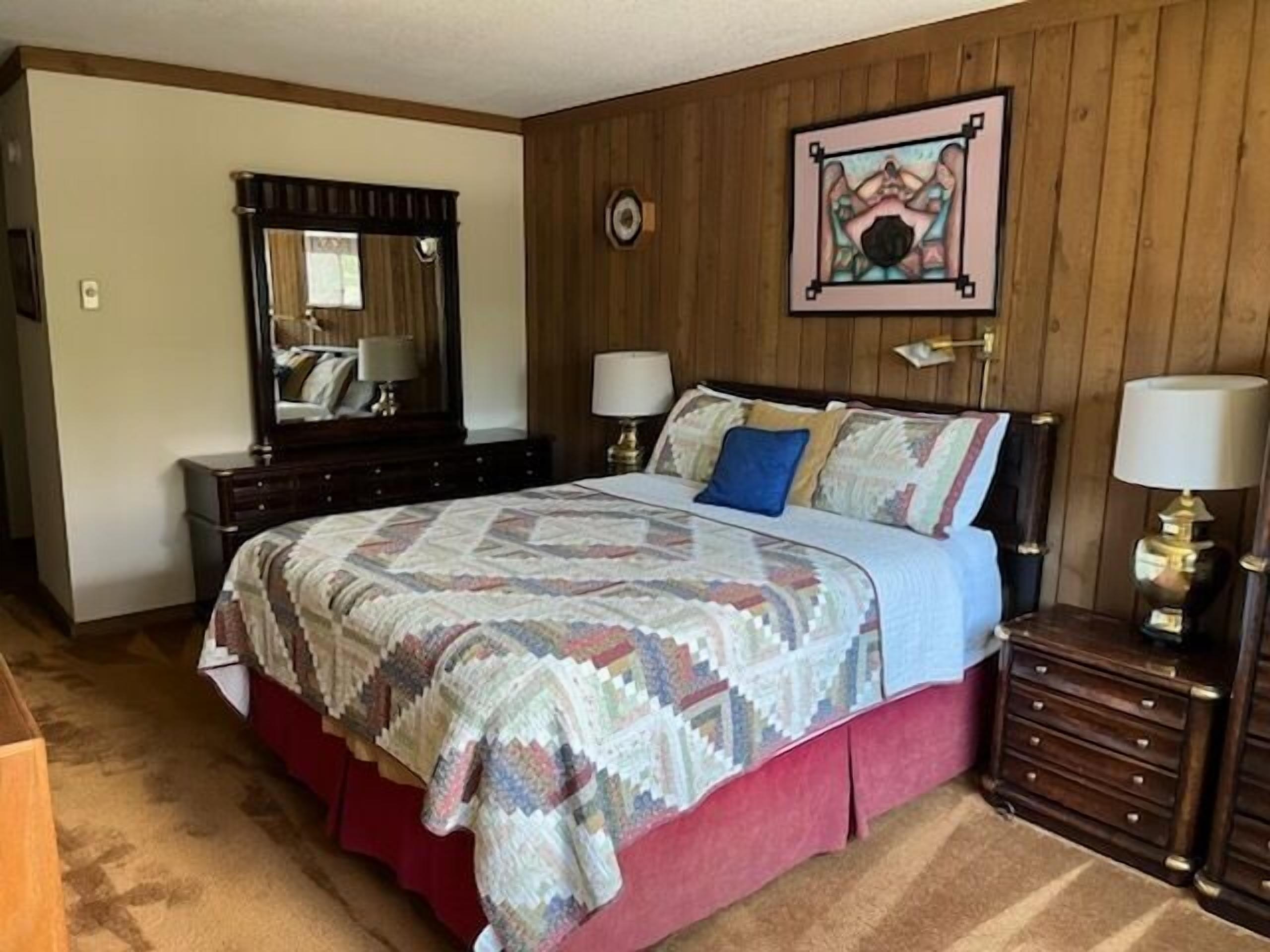 Un dormitorio con pared de madera, una cama con colcha estampada, un aparador y una mesita de noche con lámpara.