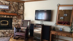 Una sala de estar con chimenea de piedra, un televisor montado en la pared, una silla de madera y un mueble de madera.