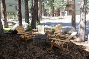 A wooden deck with Adirondack chairs and a central fire pit surrounded by trees.