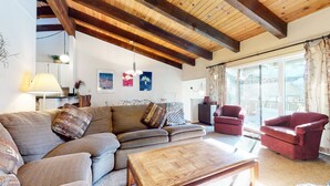 Ein gemütliches Wohnzimmer mit Holzbalkendecke, einer Sofagarnitur, einem roten Sessel und einem großen Fenster mit Vorhängen.