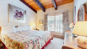 A bedroom with a floral bedspread, a wooden ceiling, a large window with curtains, a bedside table with a lamp, and a framed picture on the wall. - Spacious Home, Wood-Burning Fireplace, On Shuttle Route & Walk to Downtown (Mammoth Lakes)