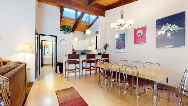 Ein modernes Wohnzimmer mit einem Holzes Esstisch, Stühlen und einer Couch. Es gibt eine Küchenzeile mit weißen Schränken und einer hängenden Pflanze.