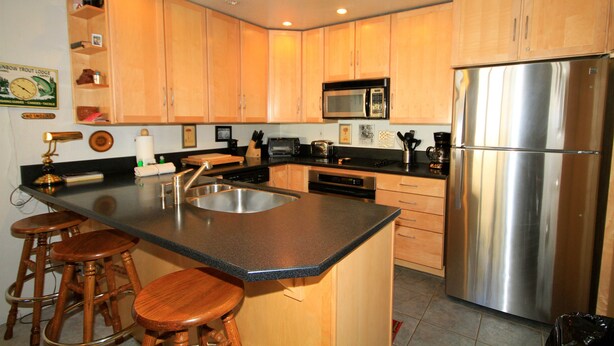 A kitchen with wooden cabinets, a stainless steel refrigerator, a microwave, and a sink. There are three wooden stools at the counter. - Courchevel #51 2 Bedroom 2 Bath (Mammoth Lakes)