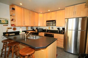 A kitchen with wooden cabinets, a stainless steel refrigerator, a microwave, and a sink. There are three wooden stools at the counter. - Courchevel #51 2 Bedroom 2 Bath (Mammoth Lakes)