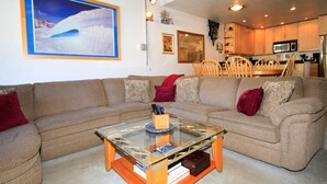 A living room with a glass coffee table, beige sofa, and red cushions. A painting of a snowy landscape is on the wall. - Courchevel #51 2 Bedroom 2 Bath (Mammoth Lakes)