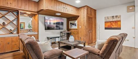 A cozy living room with wooden paneling, a large window, a fireplace, and a dining area with a table and chairs.