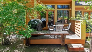 Eine Holzterrasse mit Tisch und Stühlen, umgeben von Bäumen und einem überdachten Grill.
