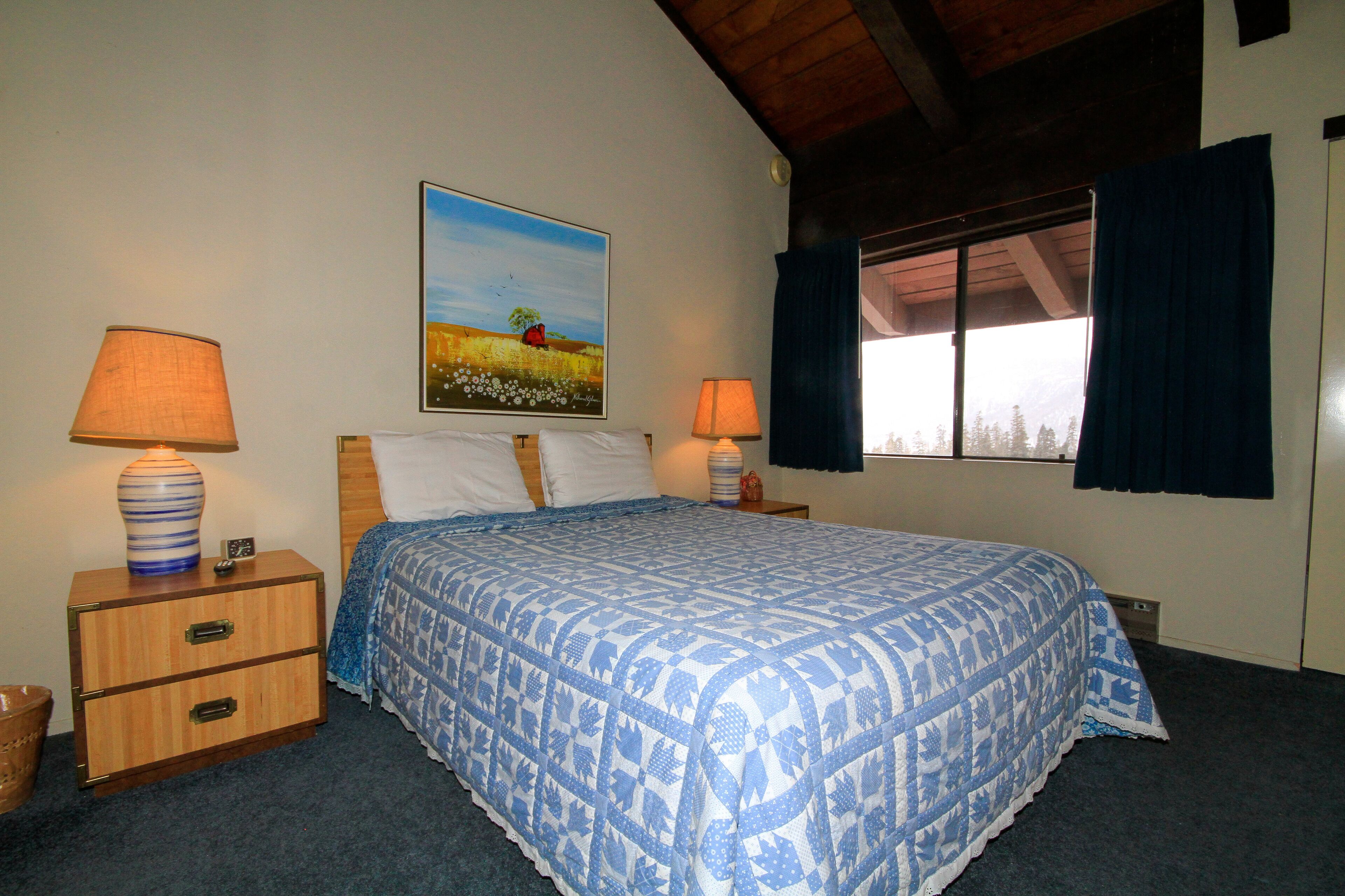 A bedroom with a bed, two bedside tables, a lamp, a painting, and a window with curtains.