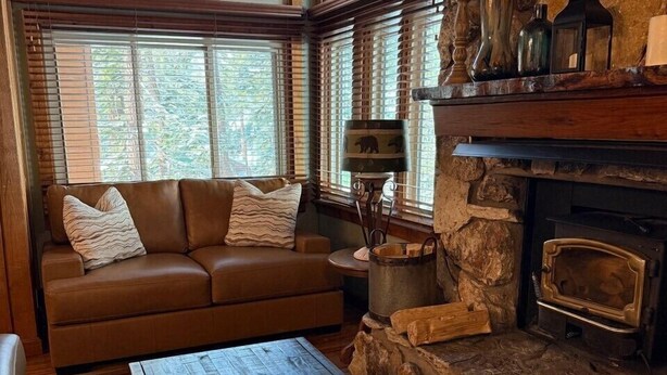 A cozy living room with a brown leather sofa, a stone fireplace, and a large window with wooden blinds. - Mountainback #79, Loft, Den (Mammoth Lakes)