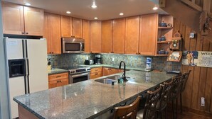 A kitchen with wooden cabinets, a granite countertop, stainless steel appliances, and a bar area with stools. - Mountainback #79, Loft, Den (Mammoth Lakes)