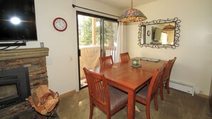 Comedor con mesa de madera, sillas, televisor, chimenea y un ventanal con vistas a árboles.