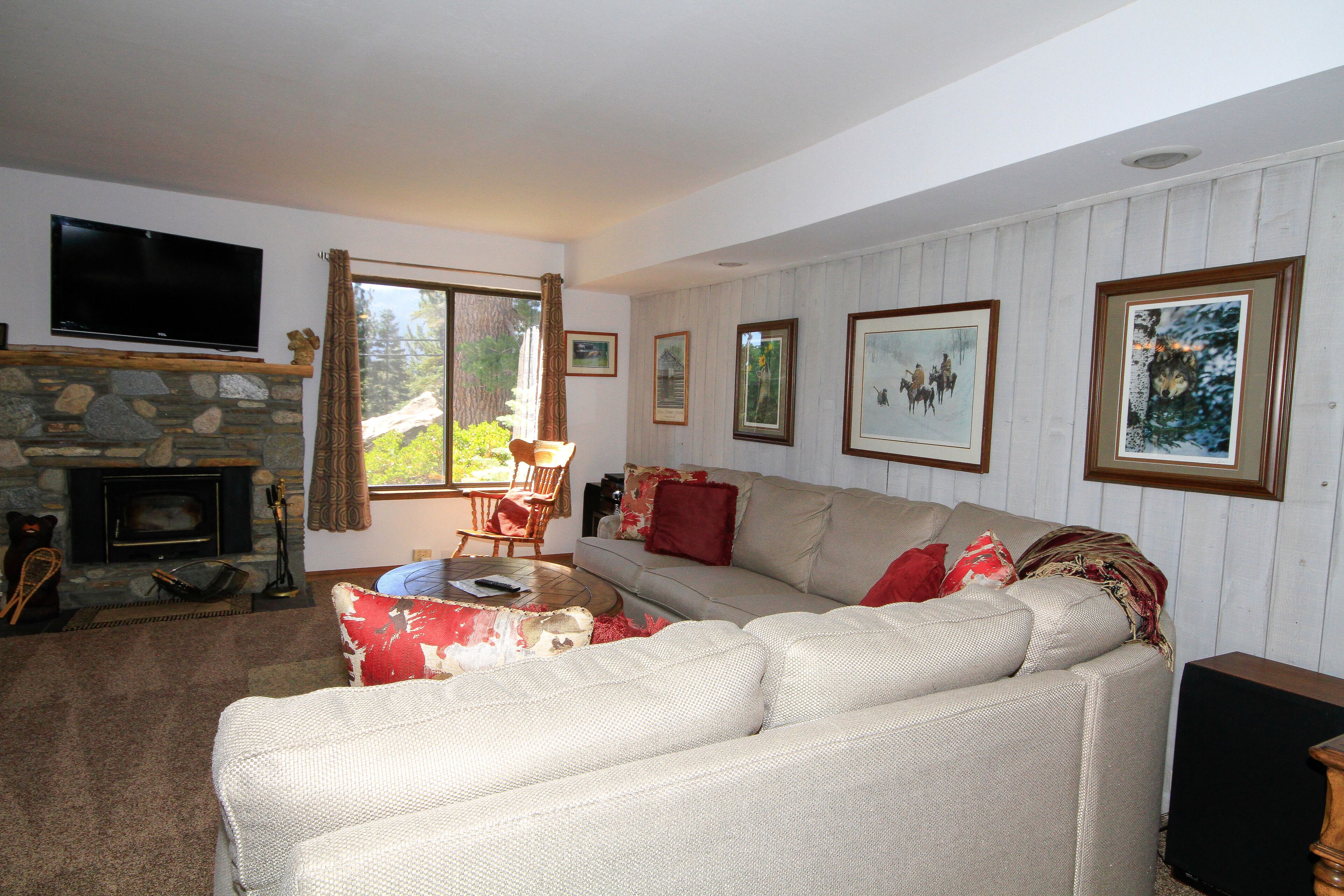 A living room with a stone fireplace, a large flat-screen TV mounted above it, a beige sofa with red cushions, and framed pictures on the wall.