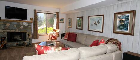 A living room with a stone fireplace, a large flat-screen TV mounted above it, a beige sofa with red cushions, and framed pictures on the wall.