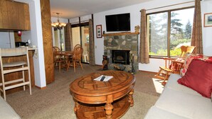 A cozy living room with a stone fireplace, wooden furniture, and large windows offering a view of the outdoors. - Discovery 4 #113 1 Bedroom 1 Bath (Mammoth Lakes)