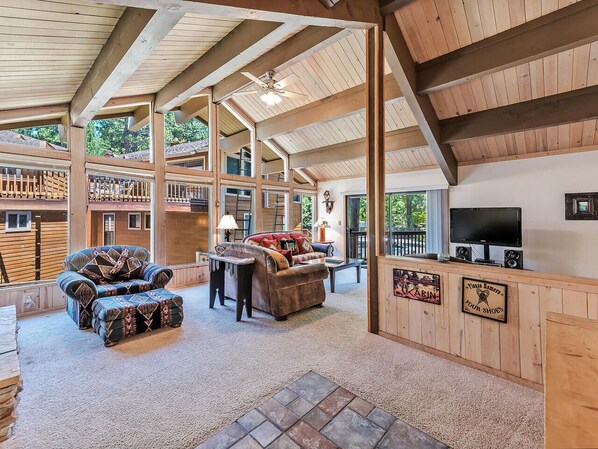 A spacious living area with a high ceiling, wooden beams, a fireplace, a television, and a sign indicating a tour shop. - 10NSE | 3BR/2.5BA Incline Village, Mtn Views & BBQ (Incline Village)