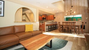 Una sala de estar moderna con un sofá de cuero marrón, una mesa de centro de madera y una zona de cocina con armarios de madera y un taburete de bar.
