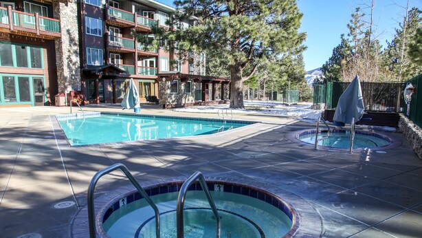 Un espace piscine comprenant un jacuzzi et une piscine classique, le tout entouré d’un bâtiment doté de balcons et offrant une vue sur la montagne.