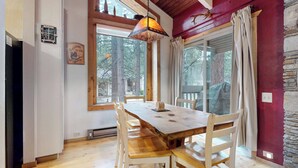Uma área de refeições com mesa e cadeiras de madeira, luminária pendente e vista para árvores por grandes janelas.