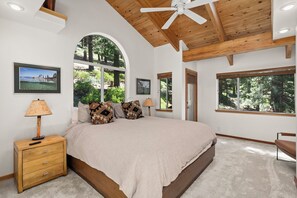 Quarto com cama grande, cabeceira de madeira, duas mesas de cabeceira com luminárias, janela com vista para árvores e ventilador de teto.