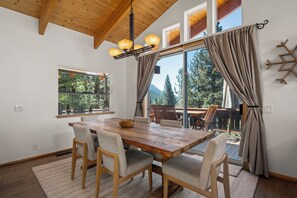 Uma área de refeições com mesa e cadeiras de madeira, janelas amplas com cortinas, e vista para árvores e montanhas.