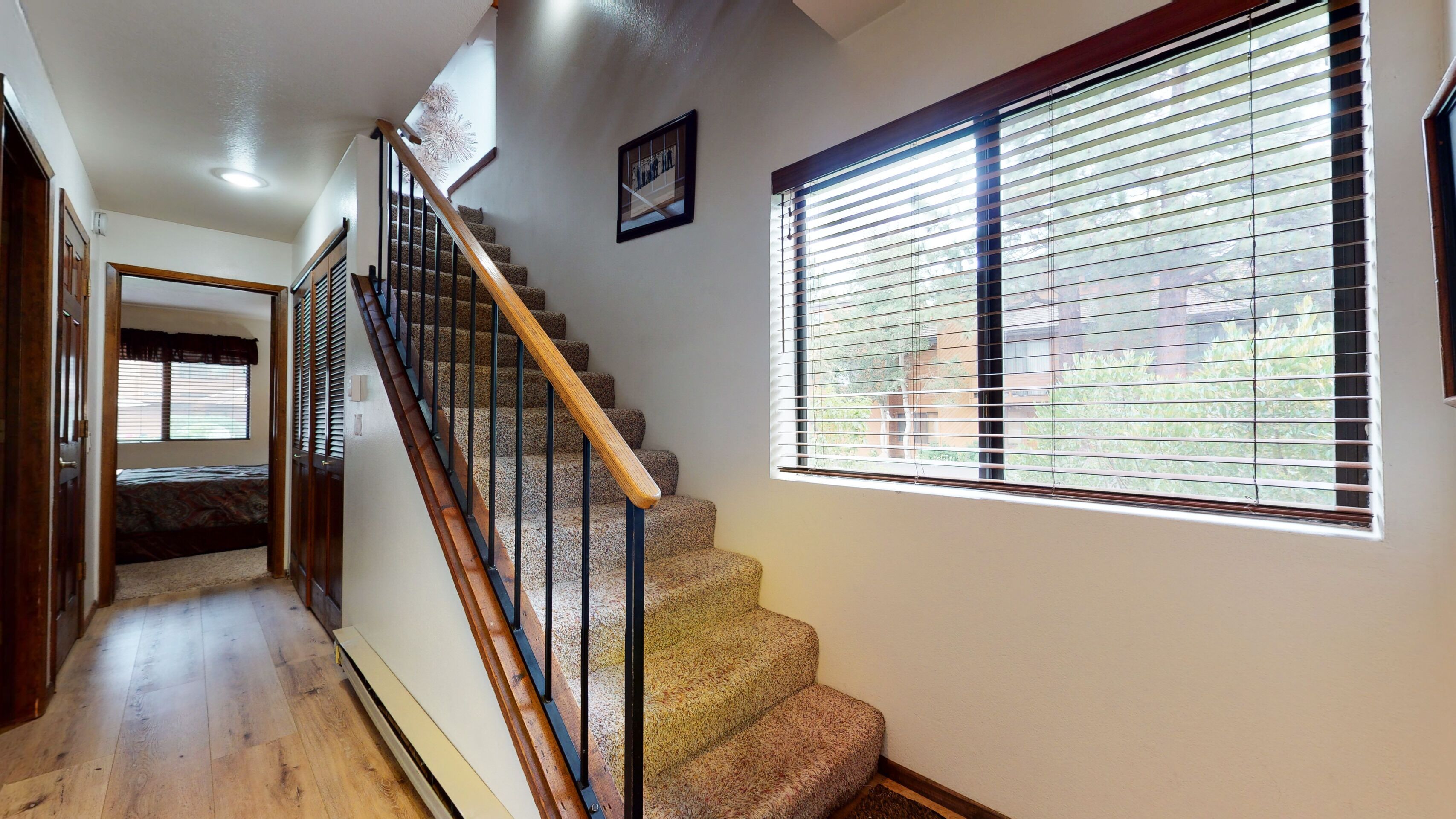 Un escalier avec des rampes en bois et des marches recouvertes de moquette, menant à une chambre dotée d’une fenêtre et de stores.