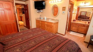 Een slaapkamer met een groot bed, houten kasten, een televisie en een badkamer met spiegel en wastafel.