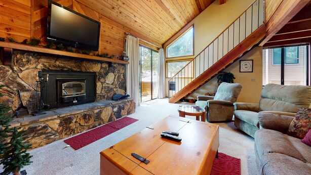 A cozy living room with a stone fireplace, a flat-screen TV mounted above it, a comfortable sofa, and a staircase leading to an upper level. - Mountain Rustic, Spacious. Washer/Dryer, On-site Pool & Near Trails/Kids Park (Mammoth Lakes)