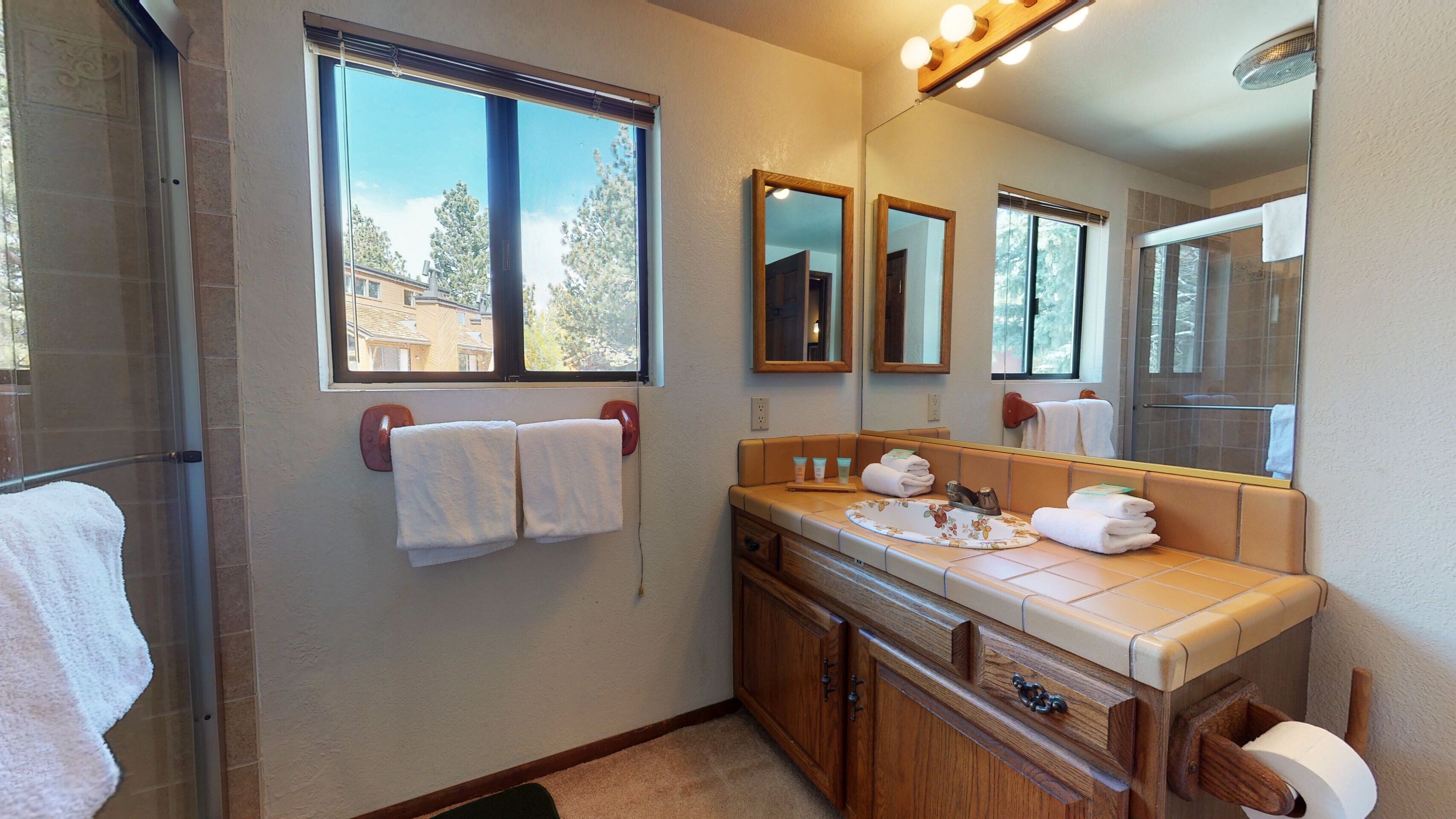 A bathroom with a shower, a vanity with a mirror, and a window with towels hanging on the wall.