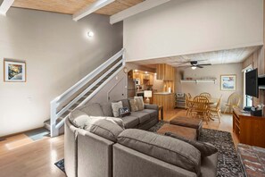 A modern living room with a gray sectional sofa, a dining area with wooden chairs, a kitchen with wooden cabinets, and a staircase with a white railing. - 214 Forest Pine | 4BR/2BA Incline Village | Pool (Incline Village)