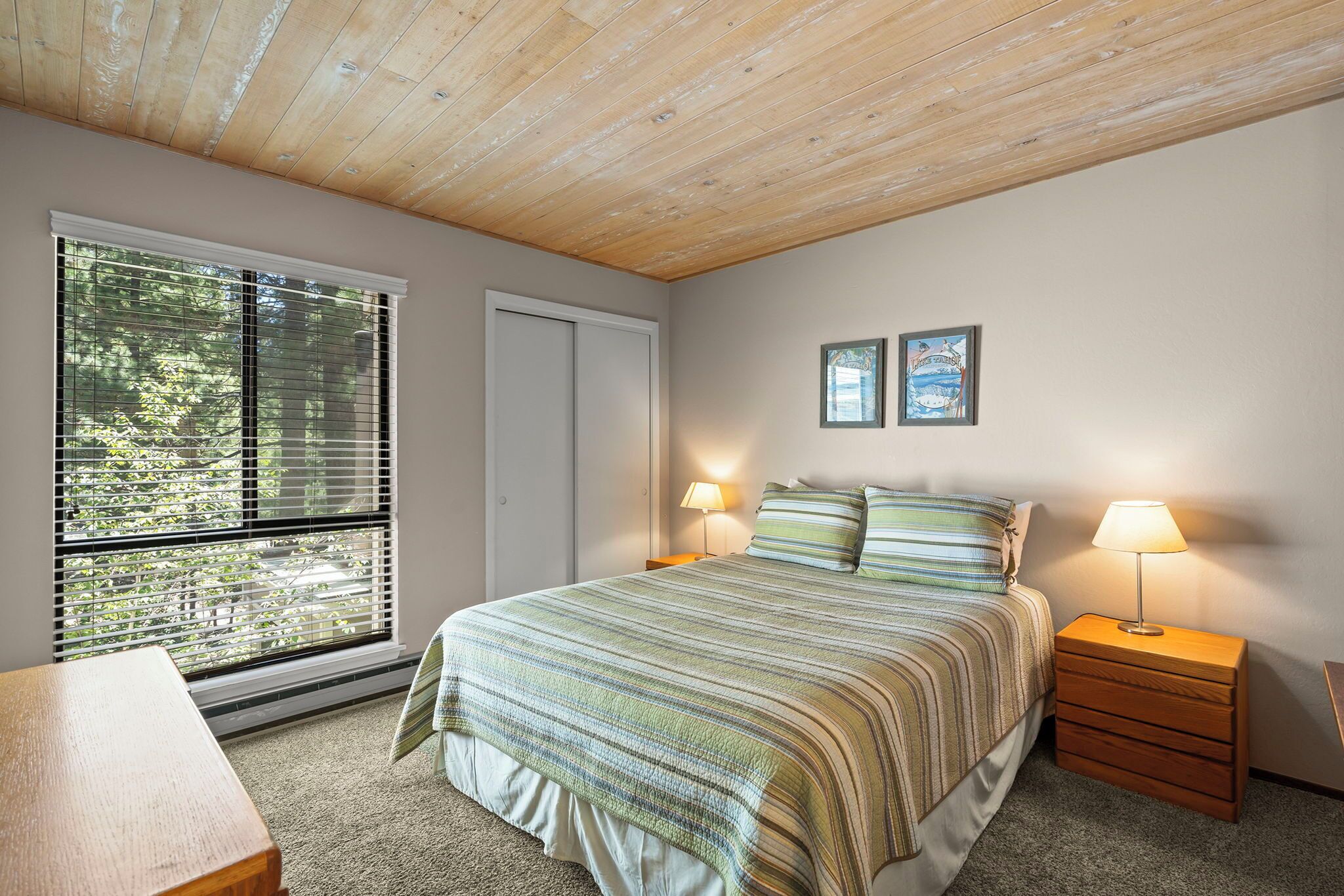 A bedroom with a bed, two bedside lamps, a wooden nightstand, a window with blinds, and a door.