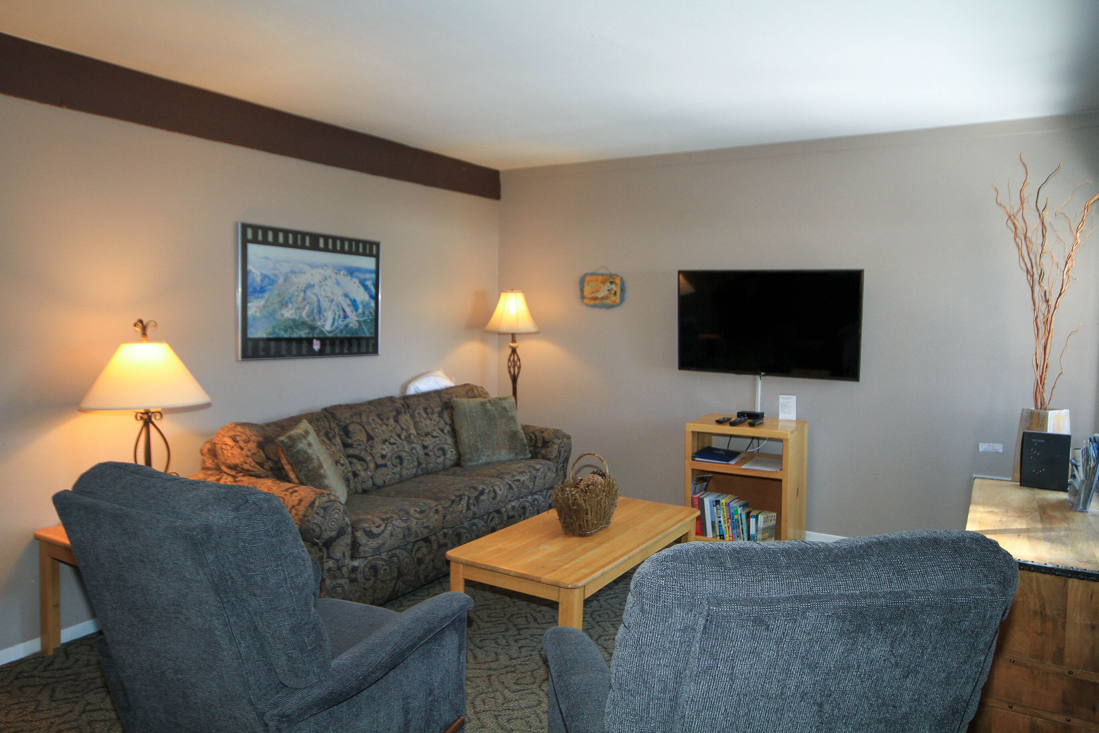 A living room with a sofa, two armchairs, a coffee table, a lamp, a TV, and a bookshelf.