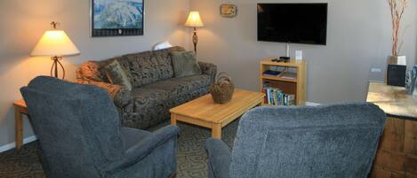 A living room with a sofa, two armchairs, a coffee table, a lamp, a TV, and a bookshelf.