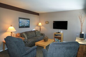 A living room with a sofa, two armchairs, a coffee table, a lamp, a TV, and a bookshelf.