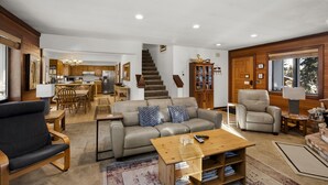 A spacious living room with a sofa, armchairs, and a coffee table. A staircase leads to a kitchen area with wooden cabinets and a dining table. - 206 Nadine | 3BR/2.5BA Incline Village | Fireplace & Patio (Incline Village)