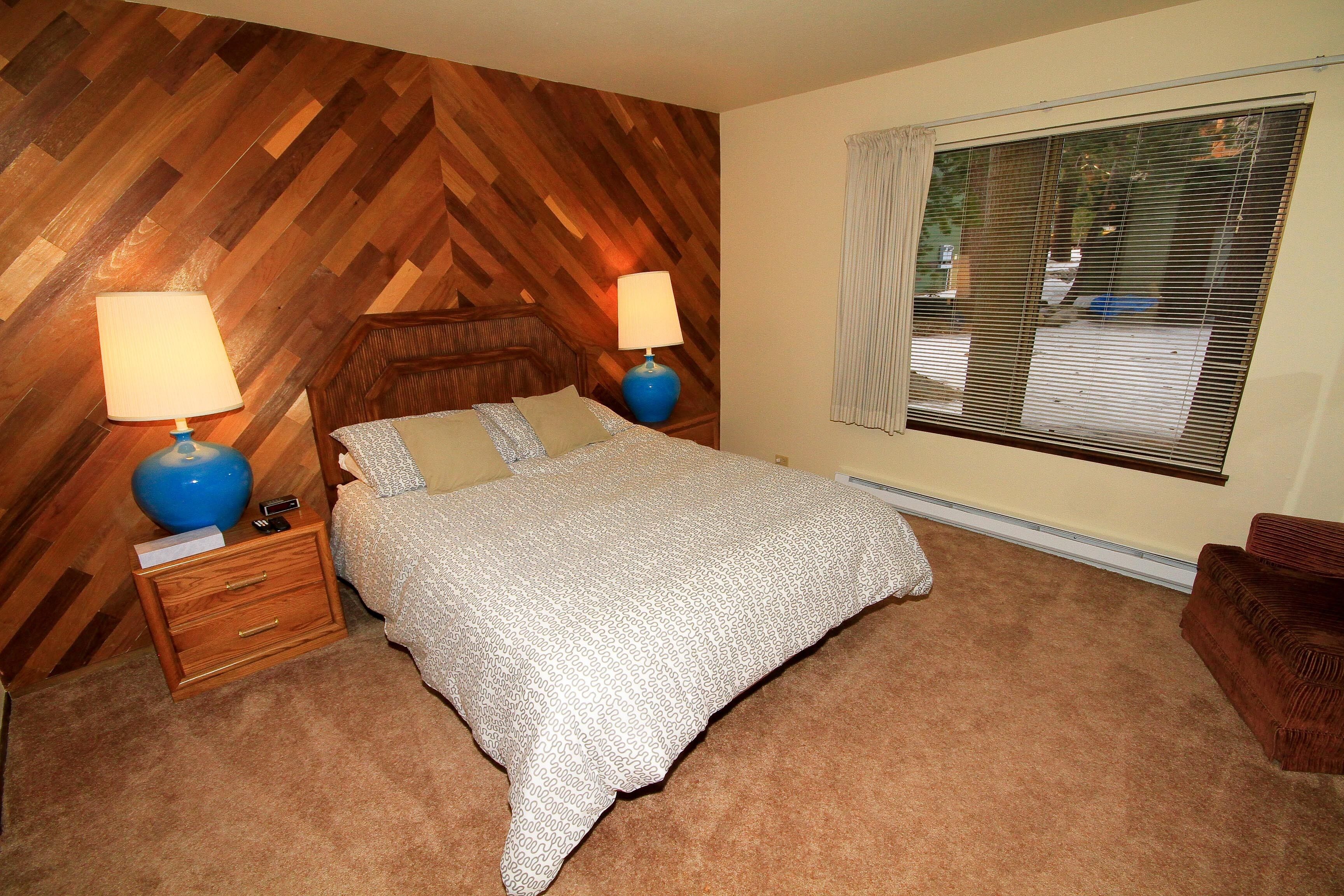 A bedroom with a wooden herringbone wall, a large window with blinds, a bed with patterned bedding, two bedside lamps, and a nightstand with a lamp and a phone.