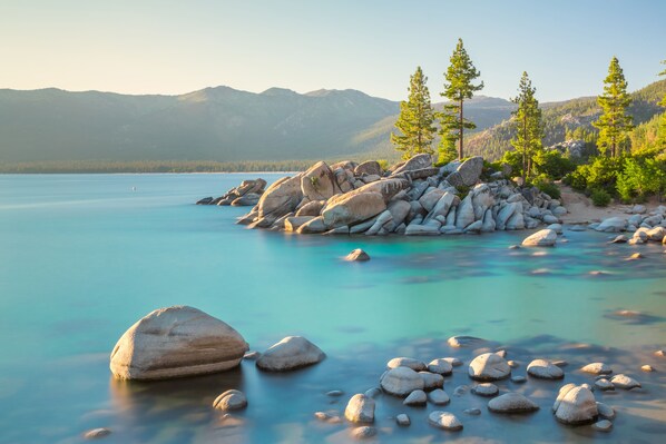 Eine friedliche Seenlandschaft mit großen Felsen, klarem türkisblauem Wasser und einem Hintergrund aus Bergen und immergrünen Bäumen.