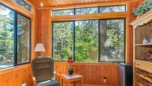 A cozy room with wooden walls, large windows, a chair, a small table, and a refrigerator.