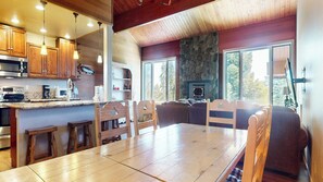 L’intérieur chaleureux d’un chalet avec une table à manger en bois, des chaises, une cheminée en pierre et un coin cuisine doté de meubles en bois et d’appareils électroménagers modernes.