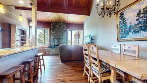 Un comedor espacioso con mobiliario de madera, chimenea de piedra y un amplio ventanal con vistas a árboles.