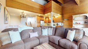 Una sala de estar acogedora con un sofá seccional, vigas de madera en el techo y una cocina al fondo.