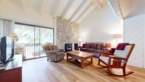 Un salon chaleureux avec un mur en pierre, une cheminée, un canapé, un fauteuil à motifs, un fauteuil à bascule et un téléviseur posé sur une table en bois.