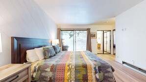 Une chambre avec un grand lit, une tête de lit en bois, une table de chevet et une porte-fenêtre coulissante donnant sur un espace extérieur enneigé.