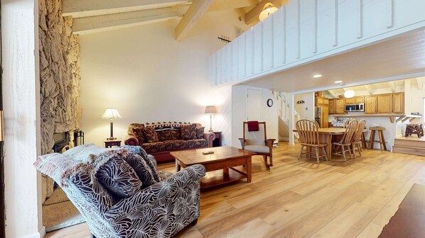 A spacious living area with a sofa, armchair, and coffee table. A kitchen is visible in the background with wooden cabinets and a dining area.