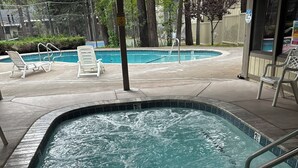 Een hottub met een houten terras eromheen, een zwembad op de achtergrond en een overdekt gebied met stoelen en een hek.