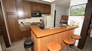 Una cocina con armarios de madera, microondas, refrigerador, cafetera y lavamanos. Hay dos taburetes en la barra.