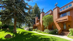 A two-story wooden building with a balcony on the second floor, a well-maintained lawn, and a large tree in the foreground. - Large, Beautiful Relaxing Location, Balcony.  Pool & Tennis Courts On-Site (Mammoth Lakes)