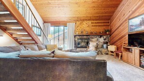 A cozy cabin interior with a wooden staircase, a stone fireplace, and a large window.